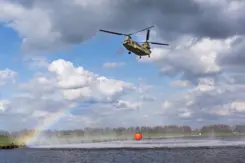 Chinook-transporthelikopter met rode bambi-bucket boven water tegen een blauwe lucht met witte wolken. Links is een regenboog te zien.