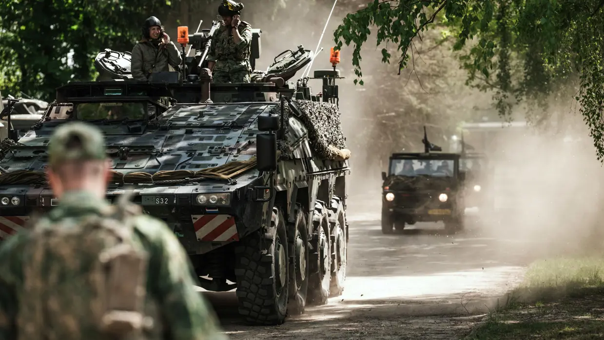 Een Boxer rijdt over een zanderig paadje, erachter rijdt een MB. Het zand stuift op. OP de voorgrond is de rug van een militair te zien.