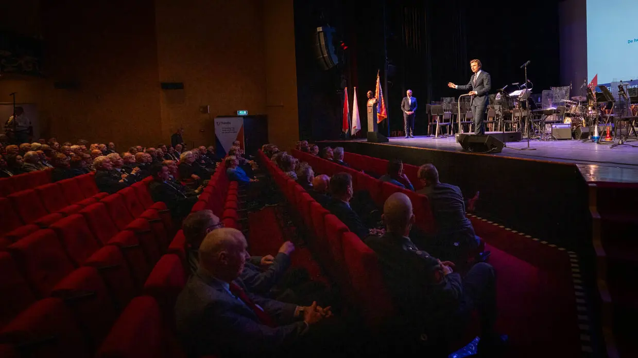 Minister Ruben Brekelmans op het podium, voor hem rode stoelen in het publiek met mensen erop.