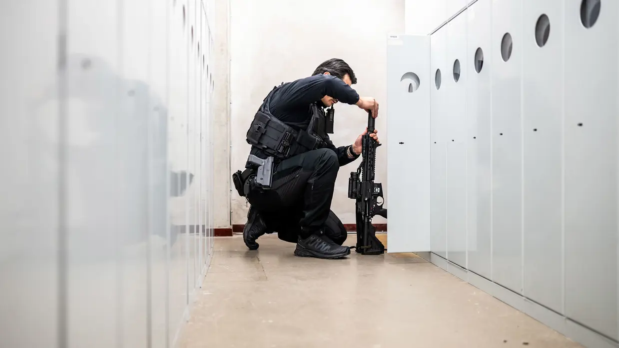 Een marechaussee knielt in een gang tussen kluisjes terwijl hij een lang wapen veilig voorbereidt voorafgaand aan de oefening. 