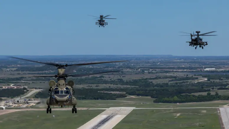 Een Chinook en twee Apaches vliegend boven een startbaan met op de achtergrond een blauwe lucht en weids uitzicht over vlak platteland.