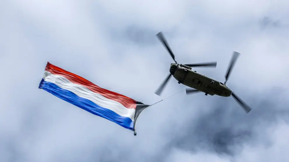 Vliegende Chinook-transporthelikopter van onderop gezien met Nederlandse vlag aan onderkant bevestigd.