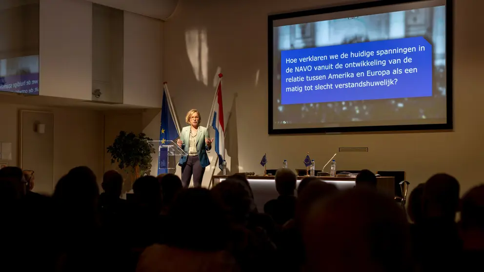 Vrouw spreekt vanaf podium volle zaal toe tijdens presentatie.