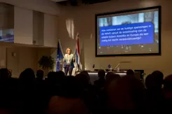 Vrouw spreekt vanaf podium volle zaal toe tijdens presentatie.