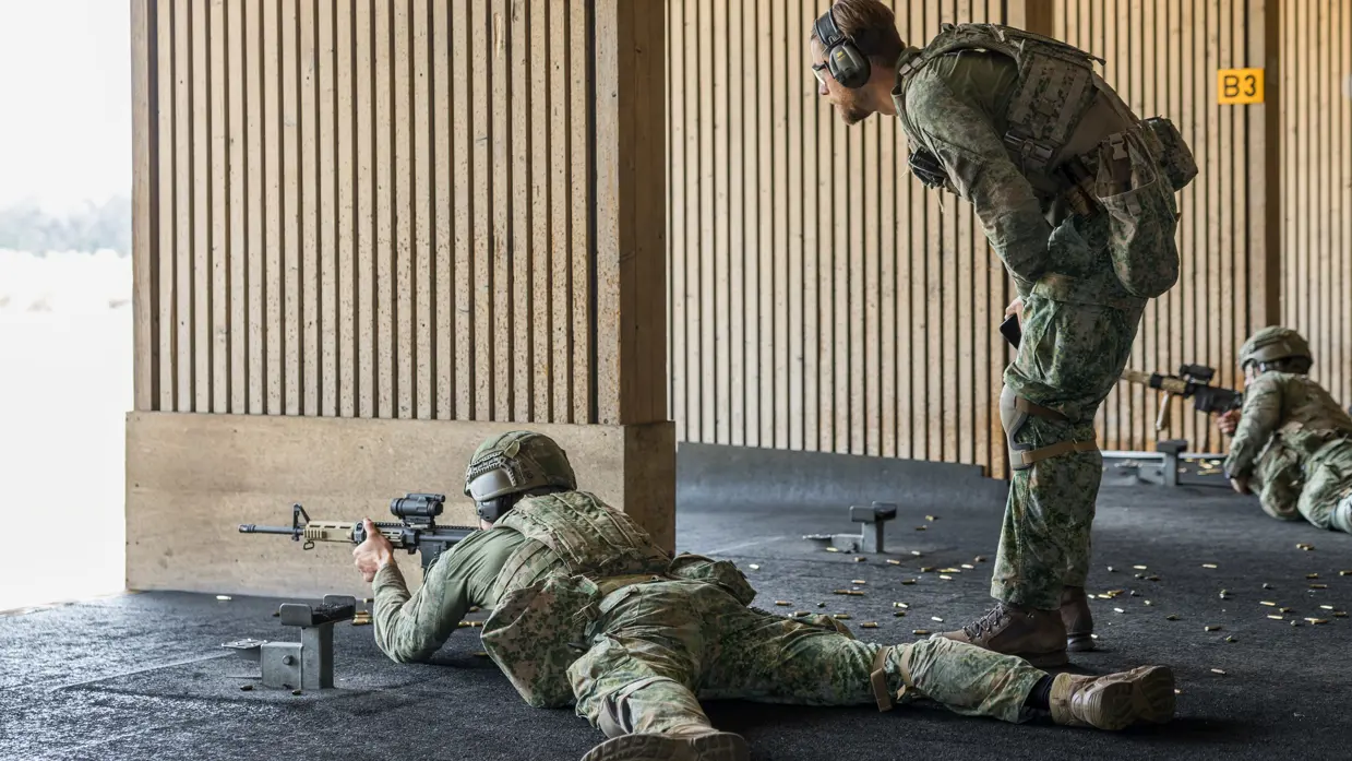 Een militair ligt op zijn buik met een wapen voor zijn gezicht op de schietbaan. Een andere militair staat achter hem en geeft hem instructies.