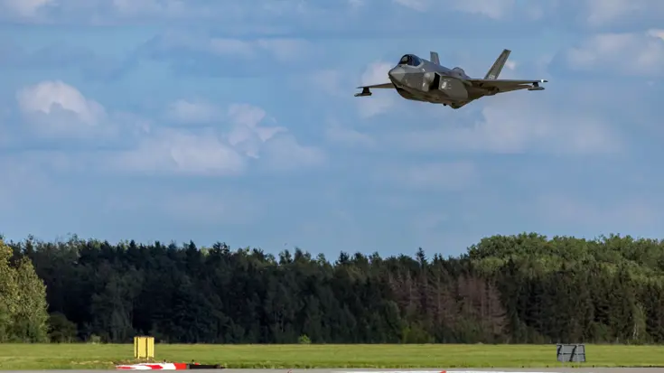 Een vliegende F-35, net boven de landingsbaan. Onder op de foto een bosrand, daarboven een blauwe lucht.