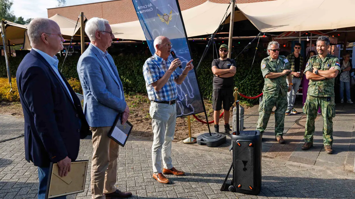 Links staan vier mannen van wie er een in een microfoon spreekt, rechts staan twee militairen.