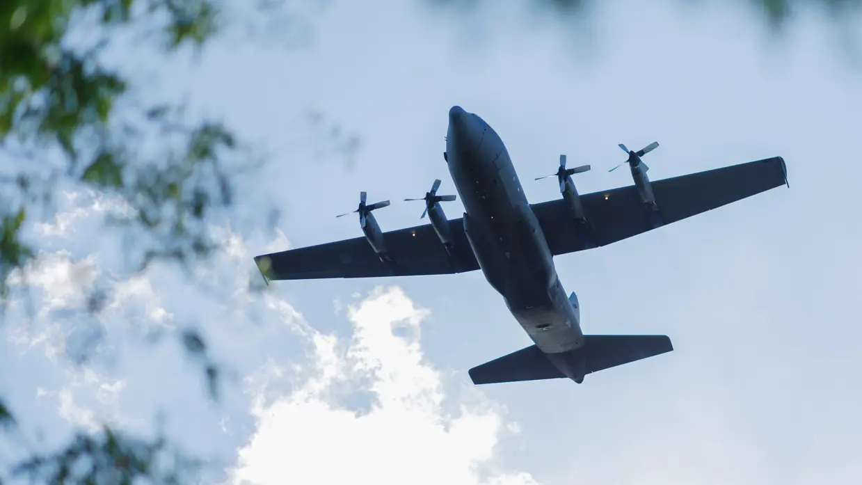 Een C-130 vliegt laag over. Links staan bomen.