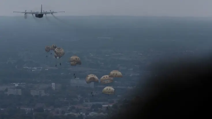 Parachutisten hangen aan hun parachutes onderweg naar beneden. Linksboven het vliegtuig waar ze net uit zijn gesprongen. 