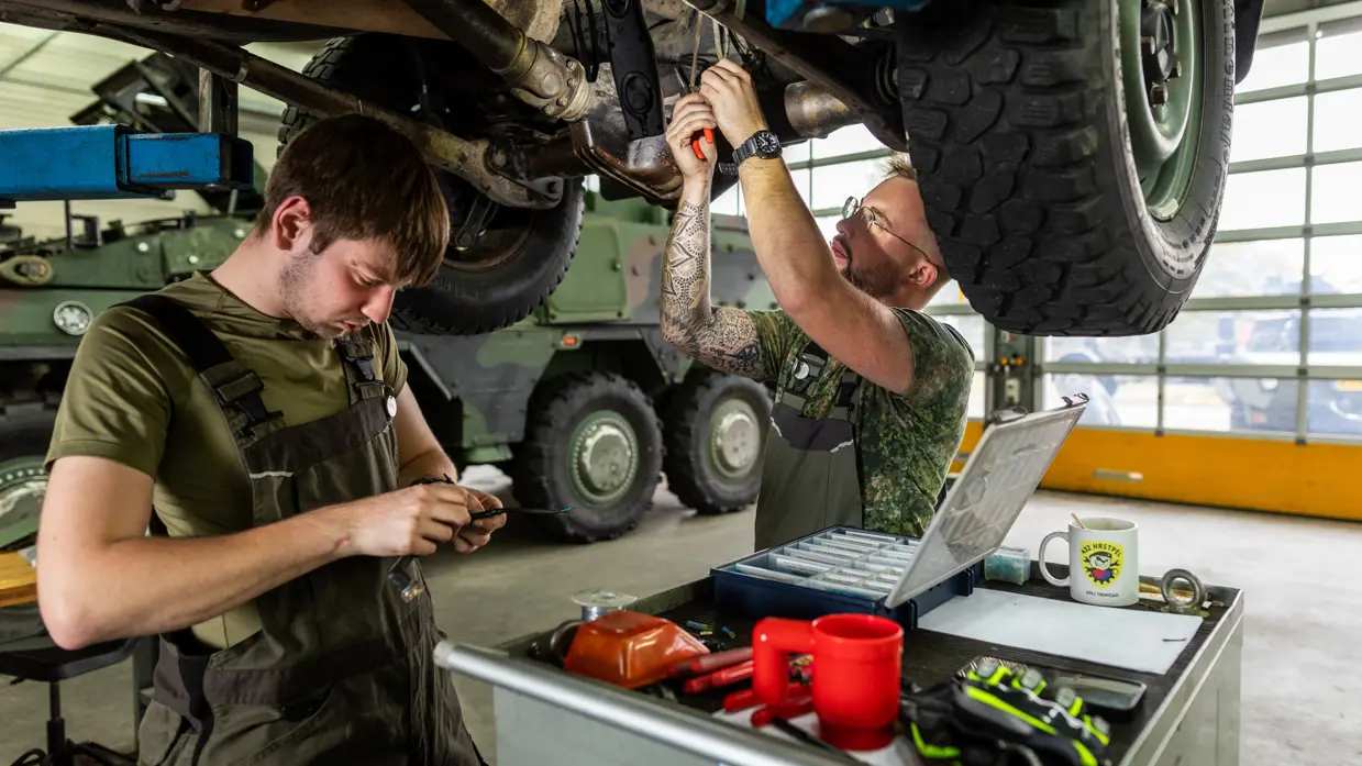 Een man in een groene overall sleutels aan een klein onderdeel in zijn handen. Een ander sleutels boven zijn hoofd aan de onderkant van een voertuig.