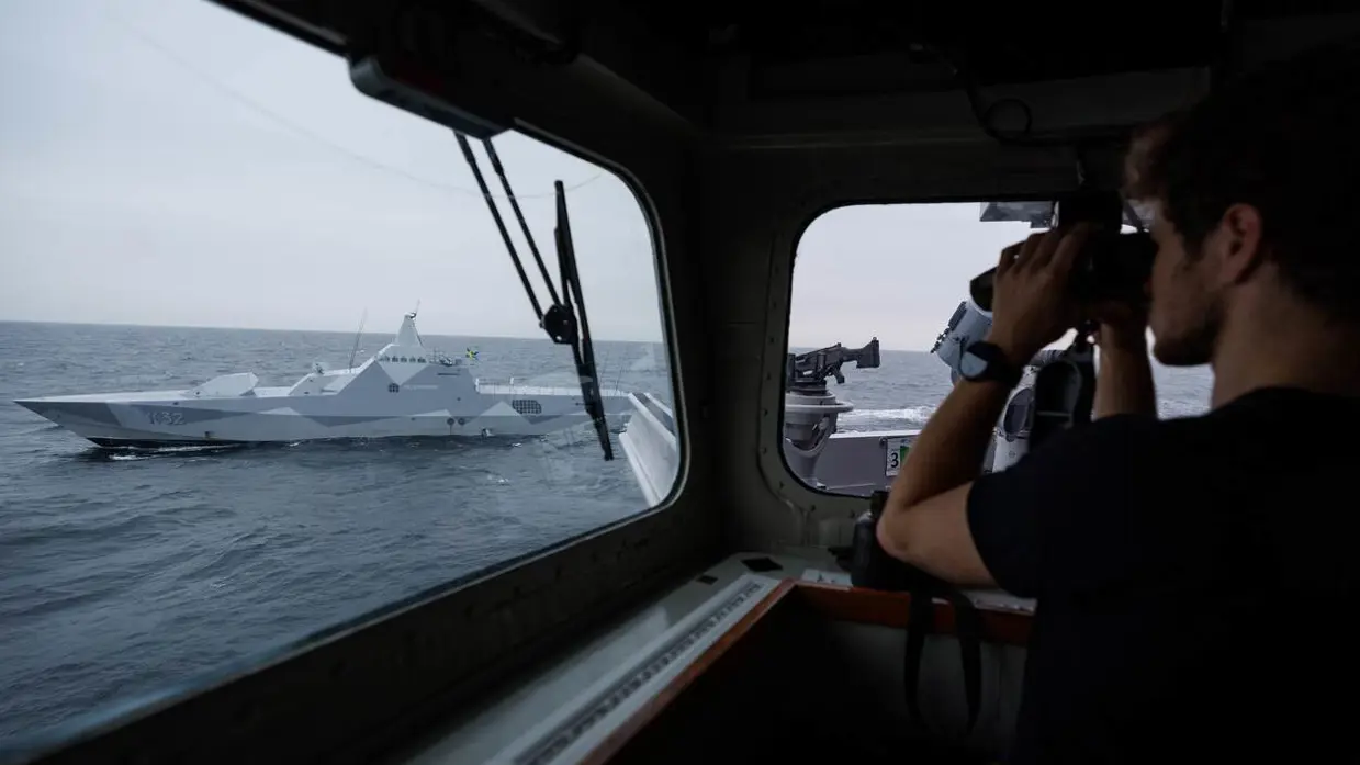 Rechts kijkt een marinecollega met een verrekijker naar een Zweeds marineschip.