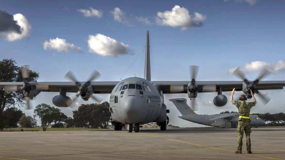 Militair op platform gebaart met armbewegingen naar een taxiënde C-130. In de achtergrond een KC-390.