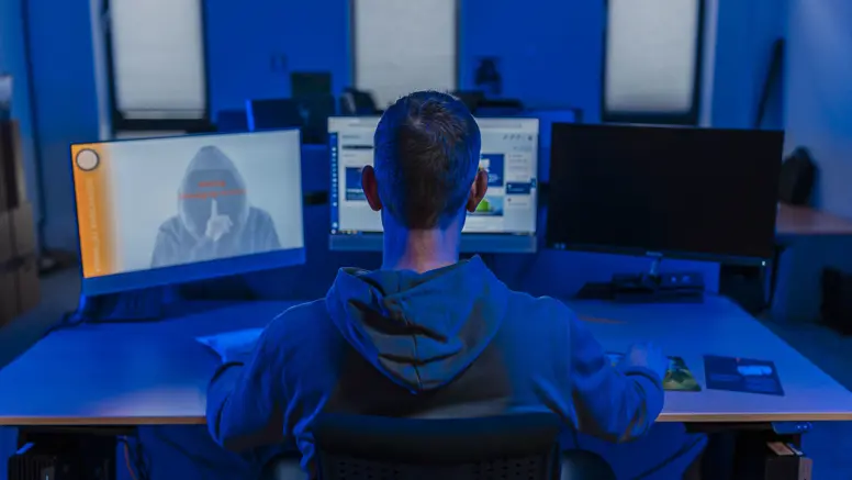 Professionele werkomgeving met een persoon aan een bureau, kijkend naar meerdere computerschermen in een blauw verlichte ruimte. Ook een silhouet van een gemaskerde figuur op één van de schermen.