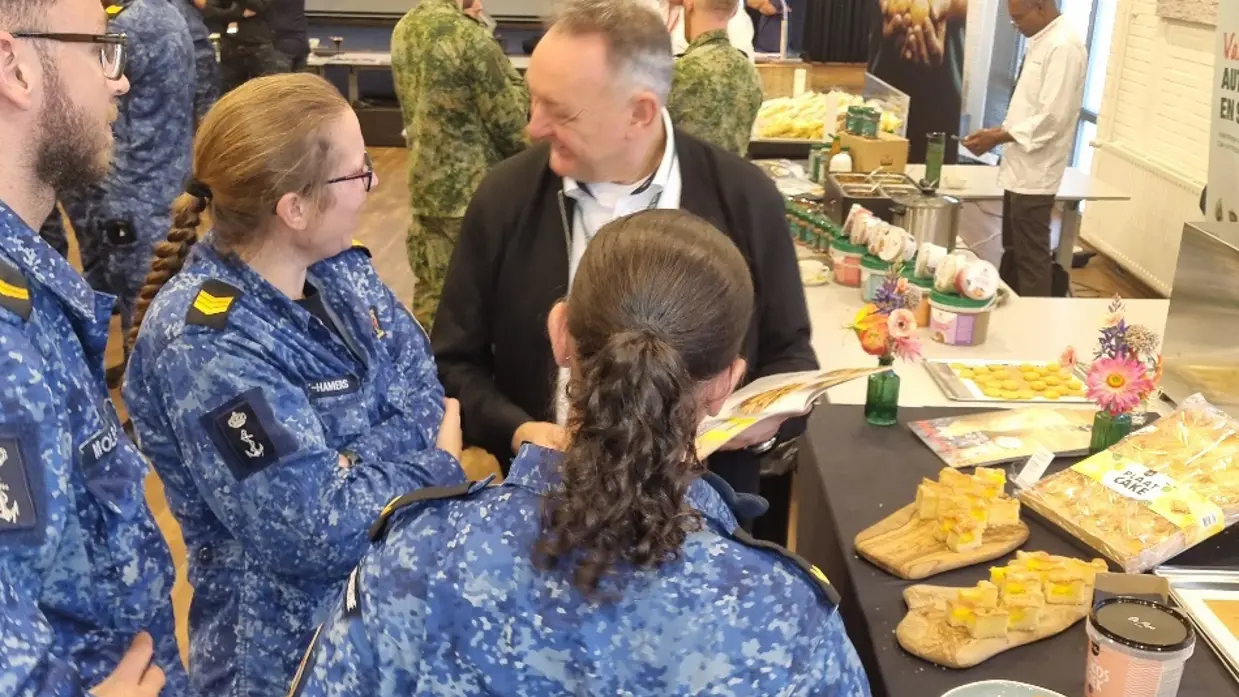 Drie personen in uniform in gesprek met een man bij een tafel voor etenswaren.
