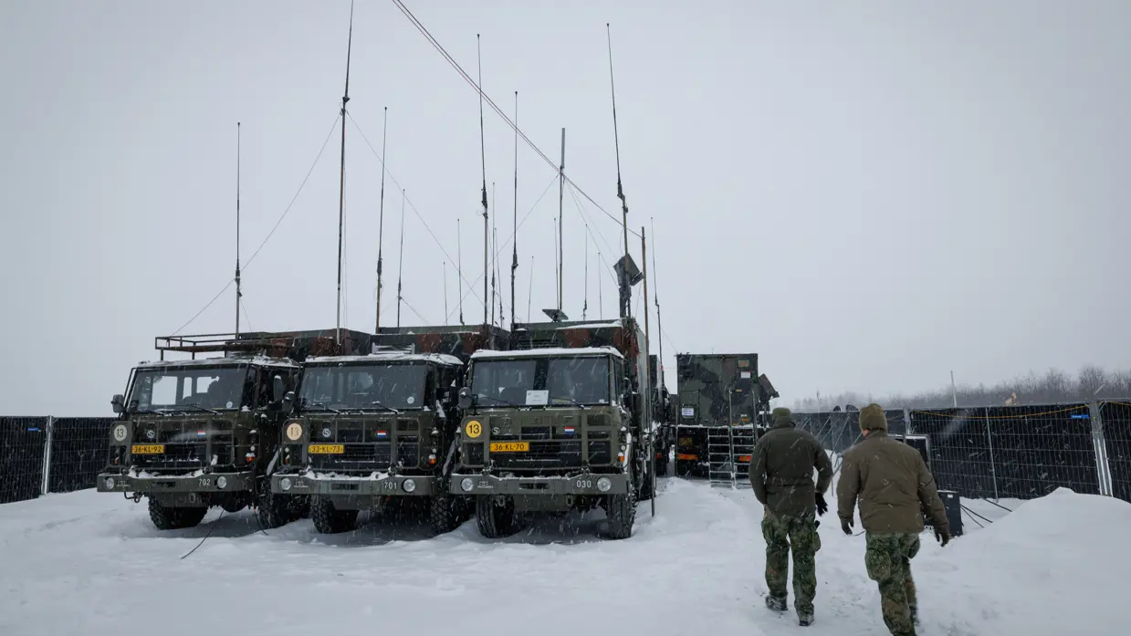 Twee militairen lopen door een sneeuwbui richting enkele groene vrachtwagens met veel antennes.