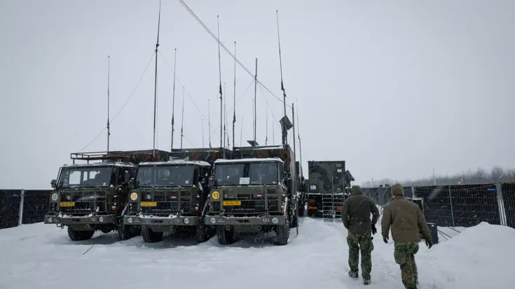 Twee militairen lopen door een sneeuwbui richting enkele groene vrachtwagens met veel antennes.