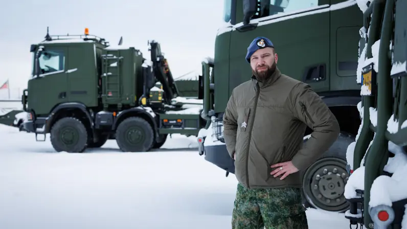 Portretfoto van Rick met zijn handen in zijn zij. Op de achtergrond enkele militaire vrachtwagens.