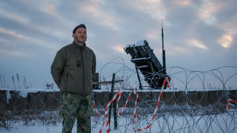 Portretfoto van Leon in besneeuwd landschap. Op de achtergrond staat de lanceerinrichting van het Patriot-wapensysteem. 