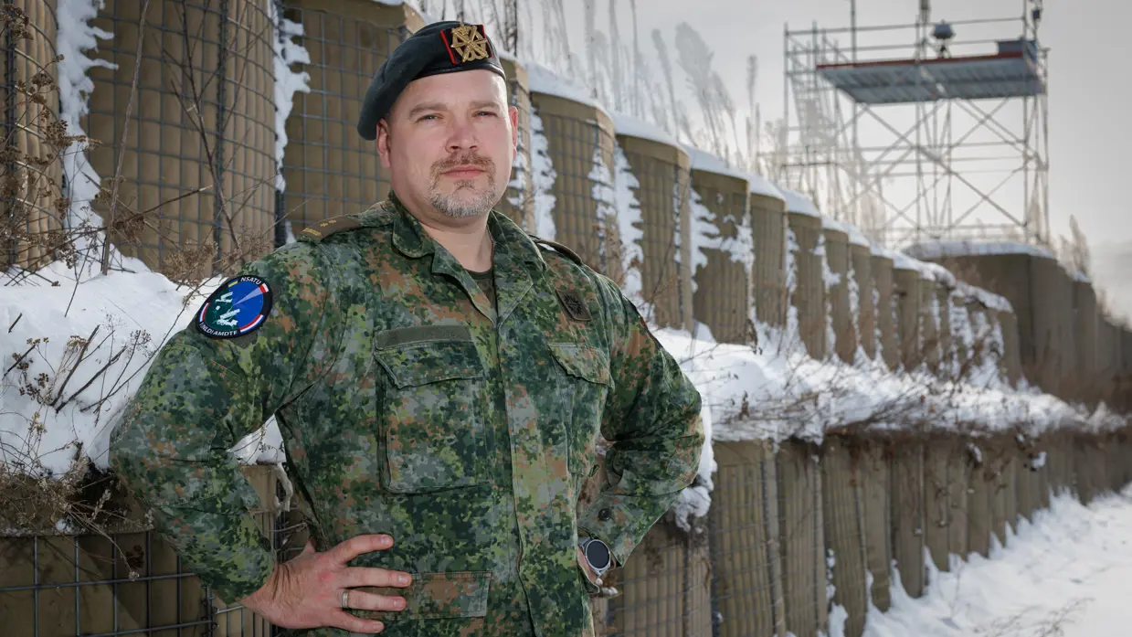 Portretfoto van commandant Dijkshoorn in gevechtstenue, met de handen in zijn zij. Op de achtergrond is een toren te zien me daarop middelen tegen kleine drones. 
