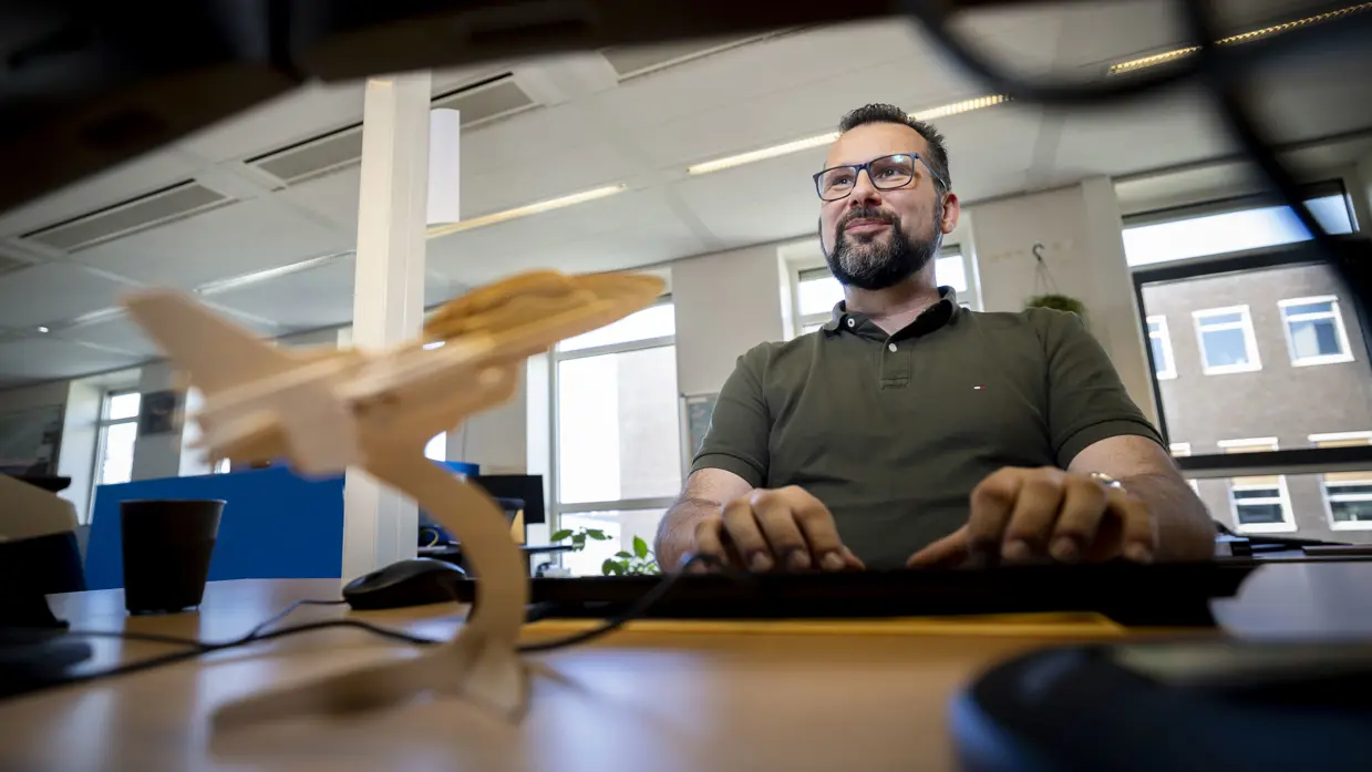 Marcel achter zijn bureau, kijkend naar zijn beeldscherm met op de voorgrond een houten vliegtuig.