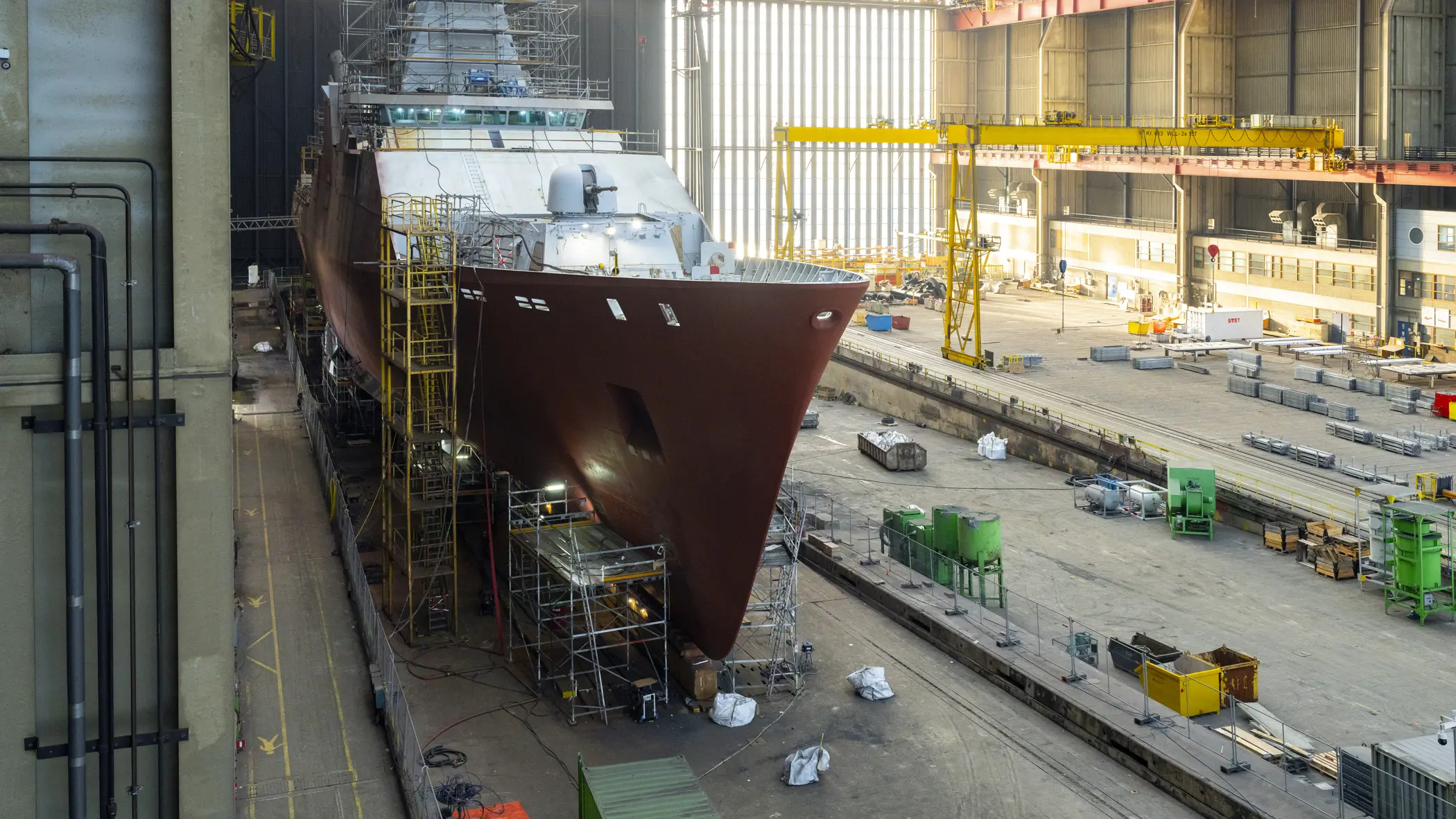 Zr.Ms. Holland in aanbouw in een overdekte scheepswerf, omringd door steigers, kranen en bouwmateriaal.