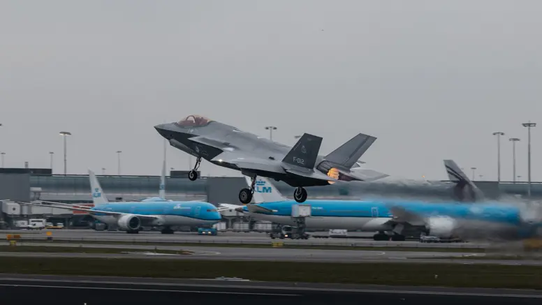 Een opstijgende F-35 op luchthaven Schiphol. Op de achtergrond zijn geparkeerde blauwe toestellen van KLM te zien.&nbsp;