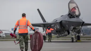 Een geparkeerde F-35 staat op luchthaven Schiphol, met de klep van de cockpit geopend. Om het toestel bewegen militairen, die onderhoud plegen. Op de voorgrond loopt iemand met een schijf met de tekst ‘remove before flight’. De luchtmachters dragen een groene militaire broek, met daarboven een oranje veiligheidshesje. Daarnaast staat een foto van taxiende F-35’s op het vliegveld. Op de achtergrond is de snelweg te zien.