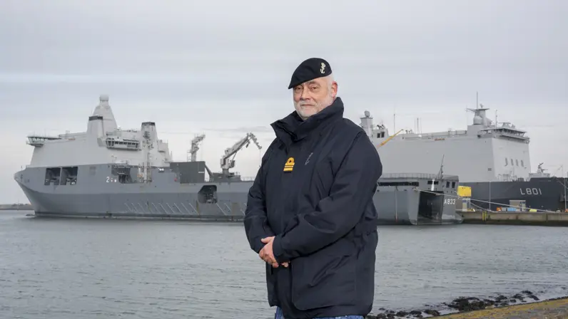 Alttekst: Een marineman poseert aan de kade voor 2 marineschepen. 