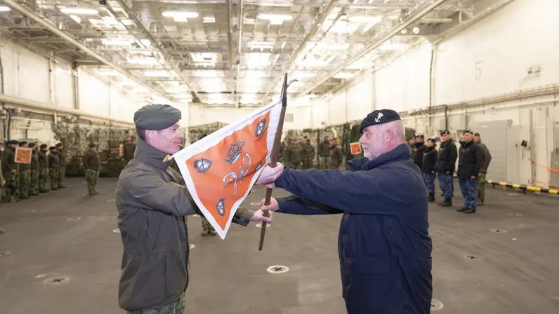 Een militair van de marine krijgt een vlag overhandigd uit handen van een militair van de landmacht.
