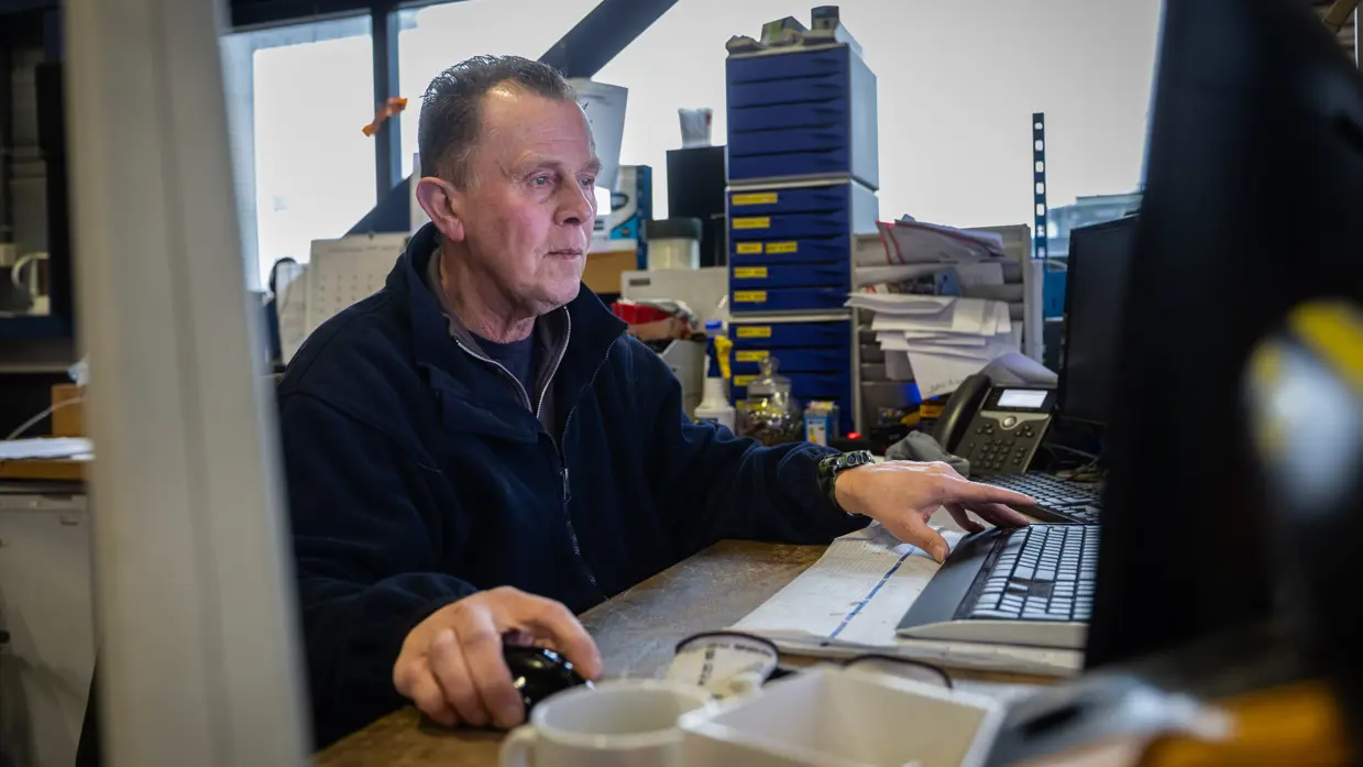 Een medewerker monitort van dichtbij hoe een pallet wordt uitgeladen. Op de tweede foto een medewerker, gezeten achter zijn bureau.