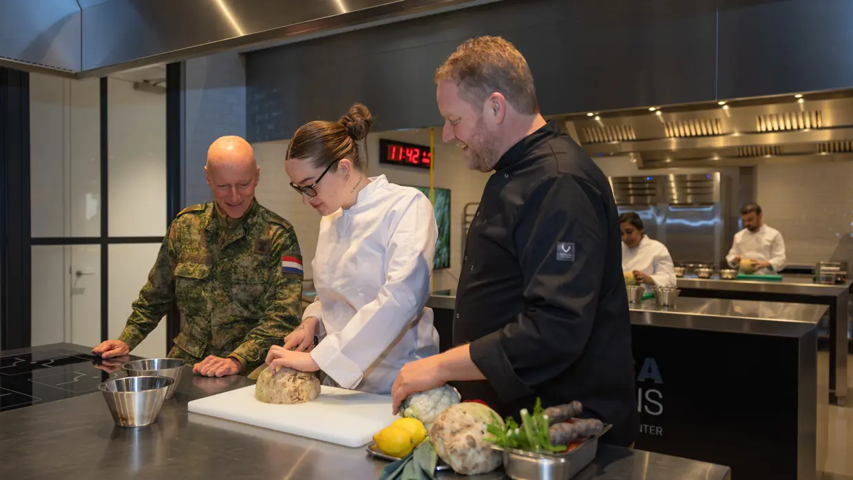 Op de linkerfoto voegen twee handen ingrediënten toe aan salades. Op de rechterfoto kijkt generaal Jobse toe hoe één van de cursisten een stuk groente snijdt. 