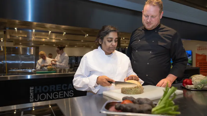 Alt: Anchila staat in een keuken en snijdt een knol onder toeziend oog van een begeleider. 