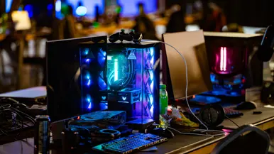 Van links naar rechts: in een zaal met veel tafels sluit een jongen zijn apparatuur aan op een computer, die lichtgevend op het bureau staat. Een man lijkt aanwijzingen te geven aan een gamer achter de computer, dan geven ze elkaar een high five.