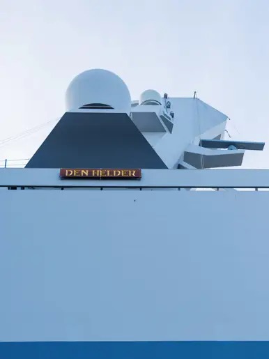 Een grijze kraan op het dek van het schip, een donkergrijze reddingsboot en het predicaat met daarop ‘Den Helder’.