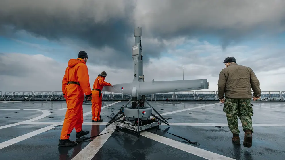 2 mannen in oranje overall en een militair zijn aan het werk met een V-BAT op het dek van een schip.