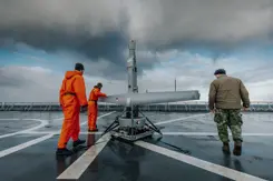 2 mannen in oranje overall en een militair zijn aan het werk met een V-BAT op het dek van een schip.