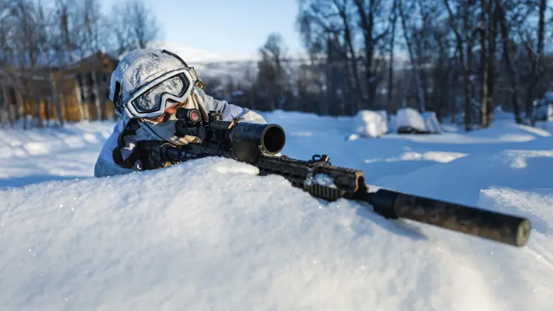 Militair in wintercamouflage ligt in de sneeuw en richt met een geweer tijdens een oefening in een besneeuwd landschap.