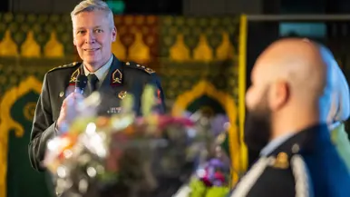  Links: Een grote, lichte ruimte waar mensen aan ronde tafels zitten te eten. Vooraan staat een man in uniform te spreken, terwijl de rest luistert. Rechts: Een glimlachende man in militair uniform met medailles spreekt in een microfoon, terwijl een andere man in uniform naar hem luistert met een boeket bloemen op tafel.