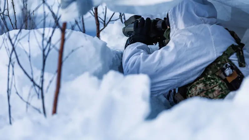 Militair in wintercamouflage observeert met een verrekijker vanuit een schuilpositie in de sneeuw.