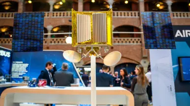 Links staat een man achter een vitrine waarin een schaalmodel is te zien van een satelliet. Op de andere foto is een schaalmodel van een andere satelliet te zien met gouden bekleding.