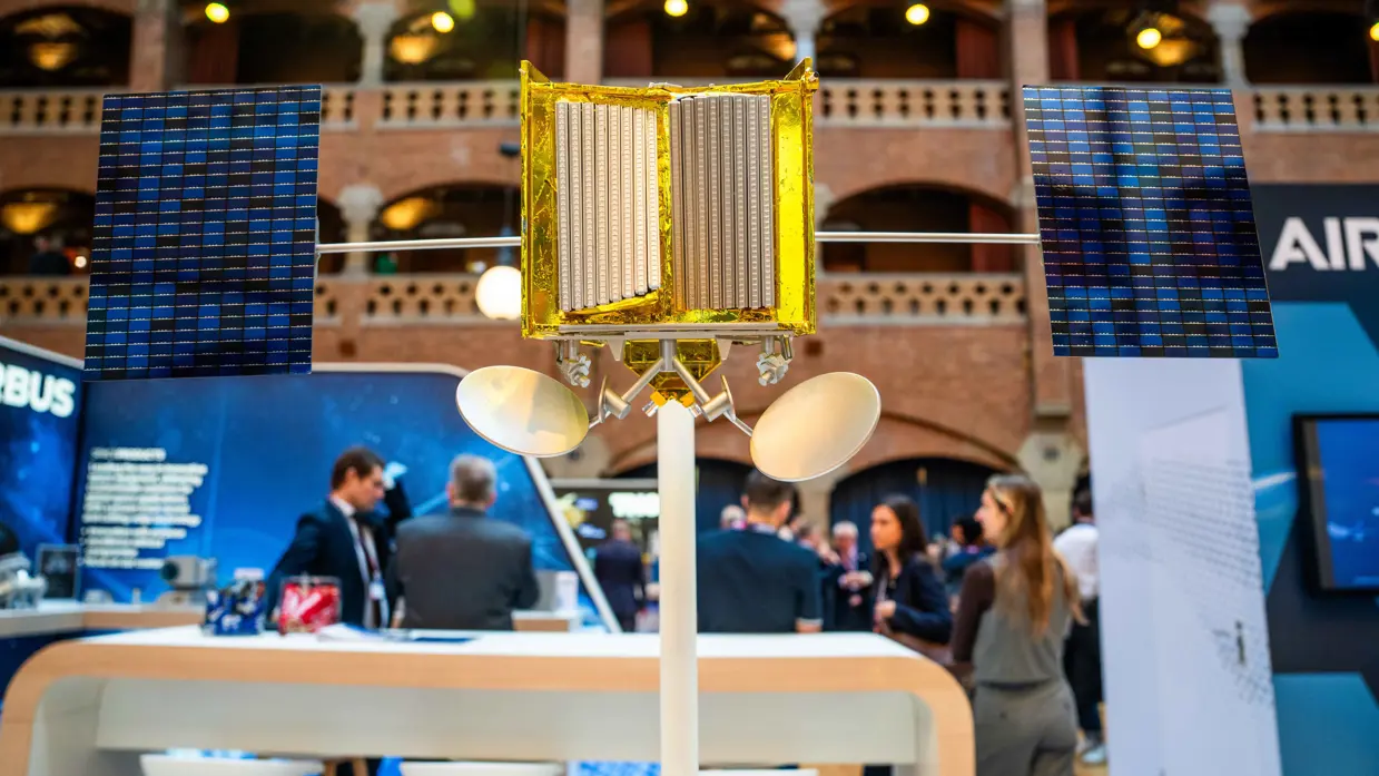 Links staat een man achter een vitrine waarin een schaalmodel is te zien van een satelliet. Op de andere foto is een schaalmodel van een andere satelliet te zien met gouden bekleding.