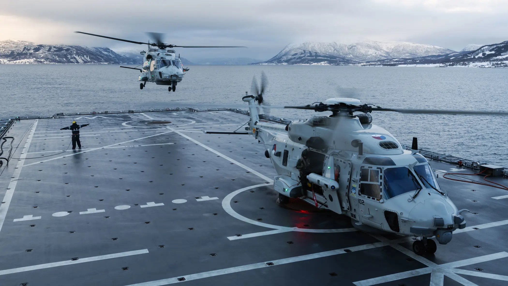 Twee NH90’s op het dek van Zr.Ms. Johan de Witt in een fjord met besneeuwde bergen; één staat op het dek terwijl de andere landt, en een vliegdekofficier geeft signalen.