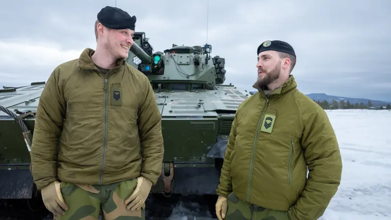Twee Noorse sergeanten staan voor een CV90 lachend met elkaar te praten.