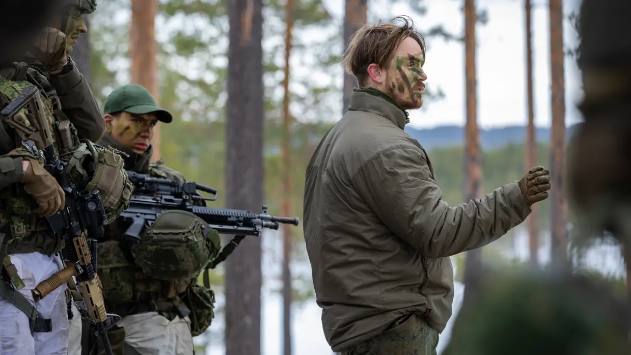 Sergeant Xander geeft in het bos in winterse omstandigheden uitleg aan collega’s. Xander heeft camouflage op zijn gezicht, achter hem staan collega’s in volle wapenrusting.