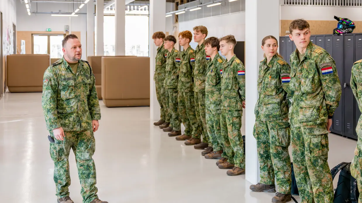 Op een strak witte vloer in een modern licht pand staan jongeren in militaire kleding op een rij. Ervoor loopt sergeant-majoor Tim Nelissen die hen toespreekt.