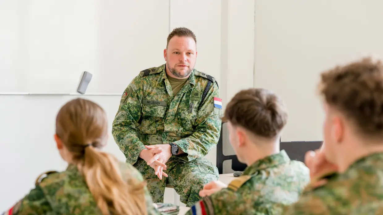 Drie jonge mensen in militair pak zitten in de schoolbanken en kijken naar sergeant-majoor Tim Nelissen die iets vertelt, zittend op de hoek van zijn bureau.