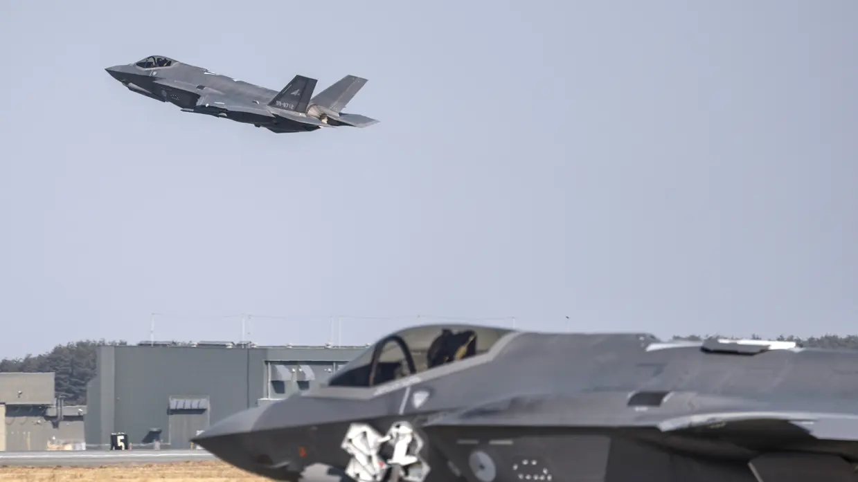 En Japanse F-35 stijgt op met in de voorgrond een Nederlandse F-35 en in de achtergrond een groen gebouw.