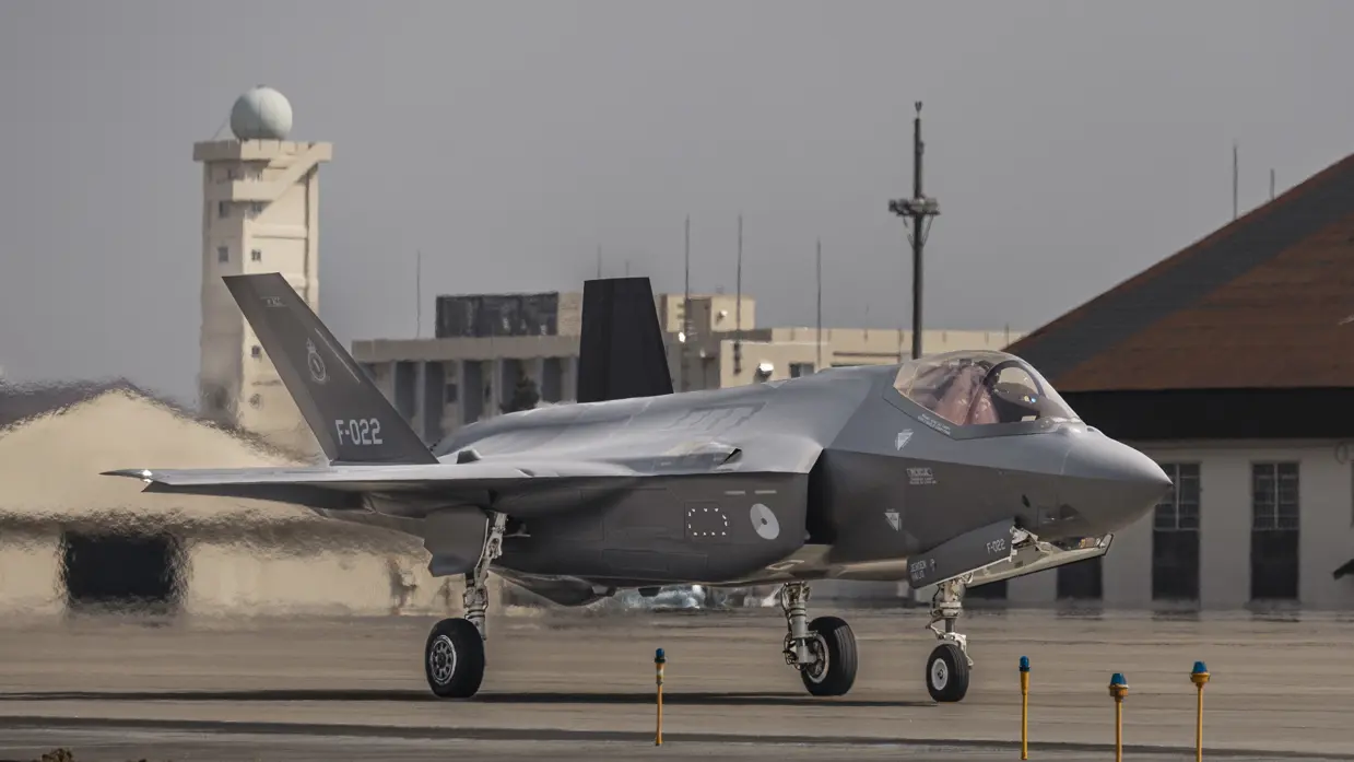 Een F-35 taxiet met in de achtergrond een radartoren, gebouwen en een hangar, en in de voorgrond gele paaltjes met daarop een blauwe lamp.