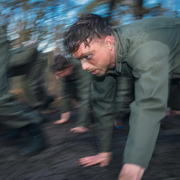 Een bezwete militair met camouflage onder het oog tijgert met collega’s door het bos.