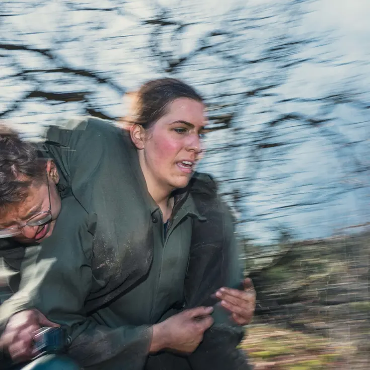 Een vrouwelijke militair rent met een bebrilde mannelijke collega in de brandweergreep.